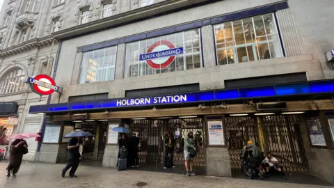 BBC/Harry Low Holborn station exterior shows closed gates and people with umbrellas up
