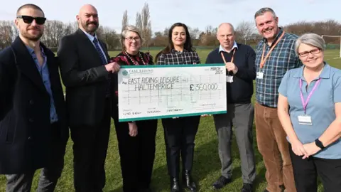 Three men and four women are standing on a football pitch with a large cheque for £360,000.