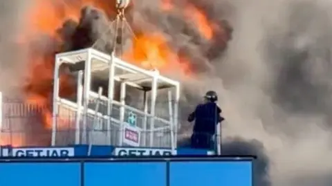 A person standing on a building. In front of him is a large fire and a white box attached to a crane