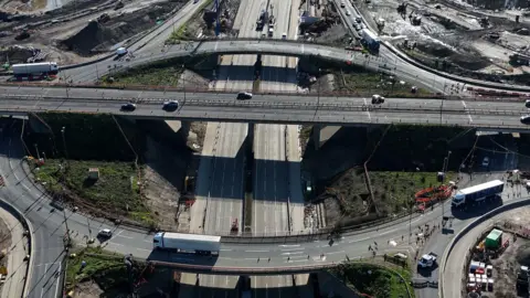 Junction 10 of the M25 seen from above. There are a number of vehicles driving on the roads, and an area of grass in the middle. 