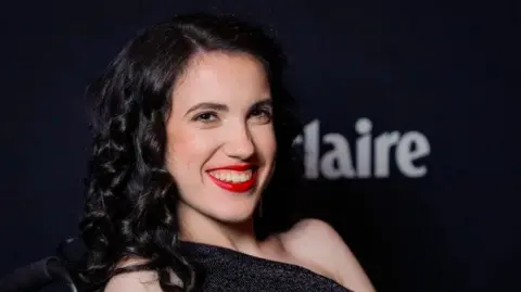 Getty Images A woman with black hair, red lipstick and a black dress smiling