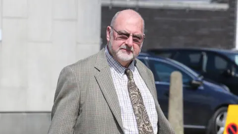 Eddie Mitchell Ifor Whittaker is looking at the camera, he is wearing a plaid shirt and paisley tie with a tweed style jacket. He wears glasses and has a short white beard