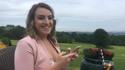 Bethan smiling at the camera with her phone in her hands. She has curled brown hair and is wearing a pink blazer and a white dress. She is sitting at a garden table with a field and trees behind her. 