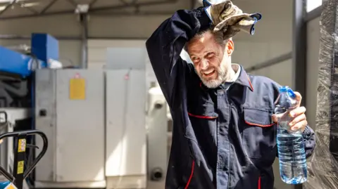 Getty Images A man in overalls wiping his forehead and holding a plastic water bottle