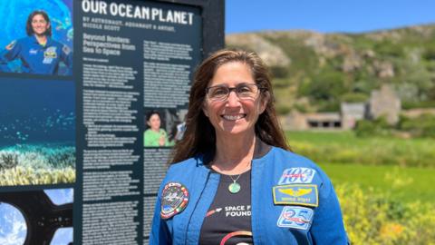 Nicole smiles at the camera next to a a sign with information about her experience in space next to her