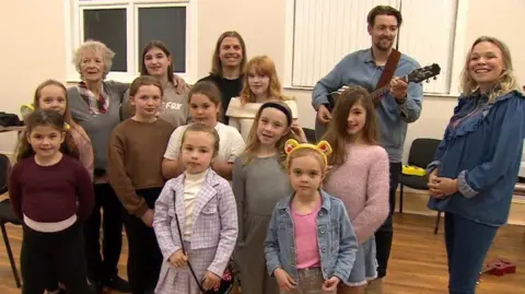 A group of 10 girls aged between seven to 13 years stand in four rows with four adults in the back row.  They are in the village hall facing the camera.  One child wears Pudsey Bear ears.  One of the adults has a guitar.