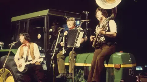 The band playing on Top of The Pops. They are holding a banjo, accordion and sousaphone, with two band members sitting on a green tractor and one standing in front holding the banjo