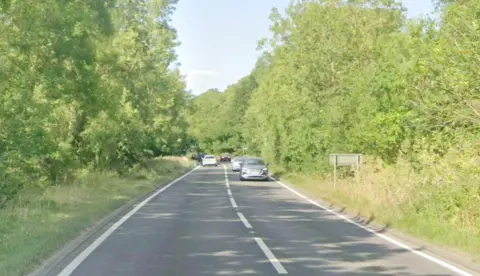 Google Google Maps images of the A509 near Little Harrowden. It shows a grey road with white dashed markings in the middle, and two solid lines at either side. The road is bordered by tall green foliage and a light blue sky can be seen at the top of the image. There are several cars on the road in both directions in the distance.
