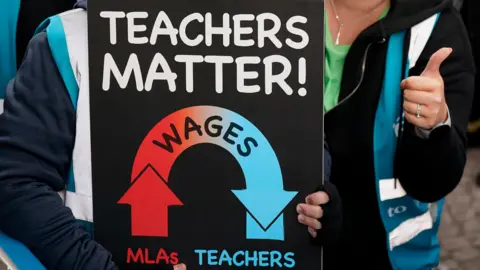 PA A close-up of a sign being held by teachers who are at a protest. The sign says 'Teachers Matter!' and shows a graphic indicating that wages for Northern Ireland assembly members are going up while teachers wages go down. Around the sign we can see the torsos of two people who are both wearing hi-visibility jackets. The person on the right is giving a thumbs up. 