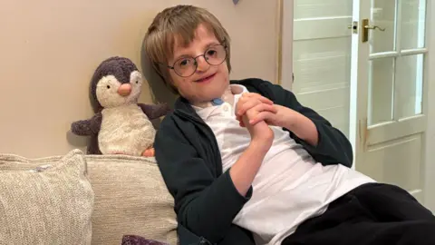 'Life-changing' surgery gives Bath boy 'first ever sleepover' 3 A boy with light-brown hair and black-rimmed glasses is leaning back on his beige sofa. He has a genetic condition and has a white plaster on his neck. He is wearing a white polo top and black fleece. He is clasping his hands together and smiling at the camera. Behind him is a grey and white toy penguin teddy.