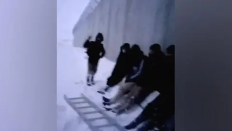 Supplied A number of migrants are seen sheltering against the wall in the snow, with a ladder at their feet - in a still from a video supplied to the BBC by a people smuggler