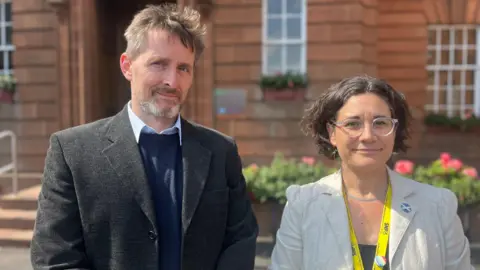 A man with greying brown hair and a beard in a dark jacket, blue jumper and shirt stands next to a woman with dark hair and glasses in a grey jacket with an SNP lanyard round her neck