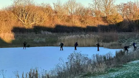 Four children attempt to cross the frozen Sherrer Pond in Swindon with others pictured nearby. The pond is surrounded by reeds, plants and trees and the grass around it is covered with ice. 