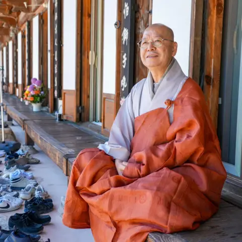 BBC/ Hosu Lee A Buddhist monk in orange and grey robes sits on the wooden porch of a traditional Korean temple building. Rows of shoes are lined up along the stone edge in front of the temple.