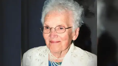 Family photo Mascha, pictured later in life. She is wearing a pale yellow jacket, pearls and a blue top. She also has metallic square glasses on, and has short grey hair. 