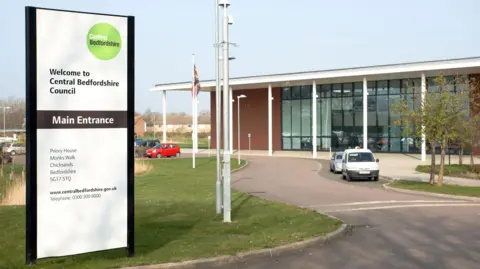 A picture of Central Bedfordshire Council Headquarters at Chicksands. The building, a modern glass-fronted construction, is in the background on the right hand side. On the left, in the foreground, is a sign saying "Welcome to Central Bedfordshire Council".