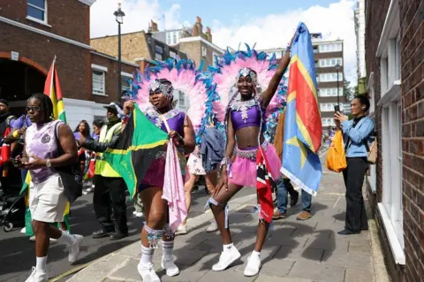 HOLLIE ADAMS / REUTERS Shauna-Leigh, 11, holds a flag of Jamaica and La'mya, 13, holds a flag of the Democratic Republic of Congo as they take part in the Children's Day Parade at Notting Hill Carnival. They are pictured posing with their flags on the street, as other people pass by. They are wearing white trainers, pink skirts, purple tops with a jewelled necklace overlaying them, and pink and blue feathered costume pieces that resemble halos around their heads.