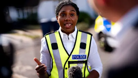 PA Media Kemi Badenoch is pictured standing in a white shirt, wearing a yellow high-vis police vest over the top. She is looking at a reporter off camera in mid-speech. Behind her is a blurred street scene.