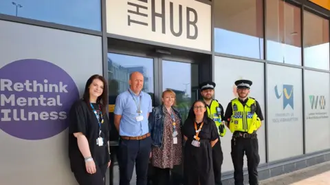 Three women and one man stand outside a building alongside two male police officers. The officers are in black outfits with black police hats and yellow fluorescent police vests. There is a sign above the door that reads "the hub".