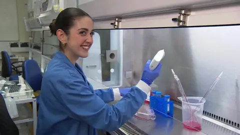 PhD student Ariana Portela working on samples in the lab. She is using a pink liquid. There are some lab containers on the surface in front of her. She is looking away, smiling. She is wearing a blue lab coat.