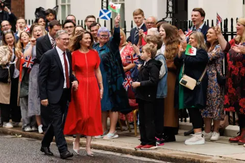 Getty Images Sir Keir and Lady Starmer walking down Downing Street having won the election. He's in a blue suit, white shirt and red tie. She's in a red dress. There are many happy-looking people with flags of the UK, Wales and Scotland.
