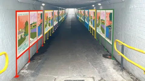 Ellie Way A long pedestrian tunnel with bright, colourful posters on both walls. The pictures show different parks and places, making the walkway feel cheerful and welcoming.