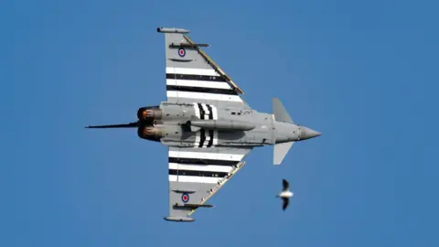 Claire Hartley A grey military plane flies against a deep blue sky. 