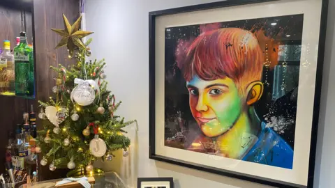 A vivid painting of a young boy in multiple colours. It is a large picture and in a frame. We can see the bar of the restaurant next to it and a mini Christmas tree. 
