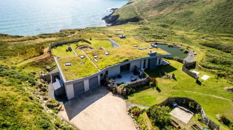 Aerial view of a modern, single-story house with a grass-covered roof nestled into a coastal hillside.