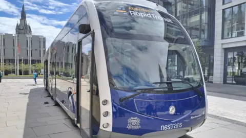 A blue and white rapid transit vehicle branded “Aberdeen Rapid Transit” is parked in the city-centre under a bright, partly cloudy sky. It is in front of the City's Town House. 