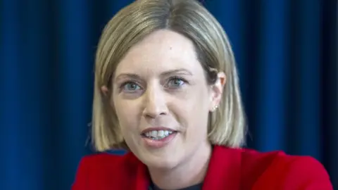 PA Media Jenny Gilruth, who has short blonde hair with a side parting, in a close-up shot in front of a blue curtain 