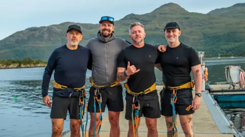 Four men in harnesses and athletic clothing stand together on a dock beside calm water, with green hills and mountains behind them.