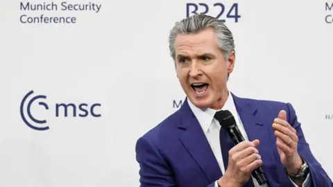 EPA California Governor Gavin Newsom in a royal blue suit and black tie speaks at a conference while holding a microphone and gesturing with his other hand