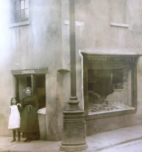 Greene family photo Black and white photo of a woman and young girl standing outside the fishmonger shop at No1 Goat Street. This business was founded in 1856