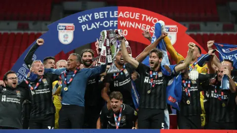 Getty Images A group of people in black striped football shirts hold up a small trophy looking happy. A red and blue banner with "Sky Bet League Two Play-Off Winner 2020" is in the background.