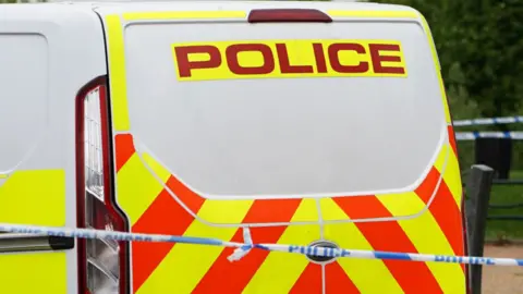 A white police van with red and yellow marking and a blue and white police tape in front