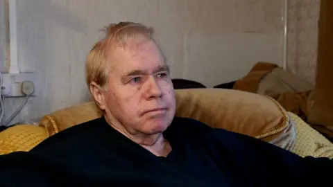 John Fairhall/BBC Jim Duell sits on a yellow sofa in a living room. He is looking away from the camera and is expressionless. He has grey hair, some of which overhands on to his forehead. He wears a dark navy blue jumper. 