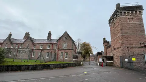 The barracks is a group of traditional, brick-built buildings with long narrow windows with white frames. The complex is surrounded by a high, wire fence.