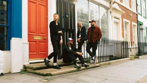 As It Is An artistic picture of a band. They are standing on the steps of some houses with colourful doors. 