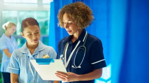 Nursing sister supervises a young ward nurse standing at the foot of a hospital bed chatting about the medical chart that she is holding . In the background a young nurse checks the dressing pack that she has got out of the trolley .