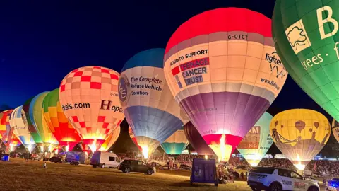 A number of balloons are tethered to the ground in the night. They are lit up, showing their different colours and designs.