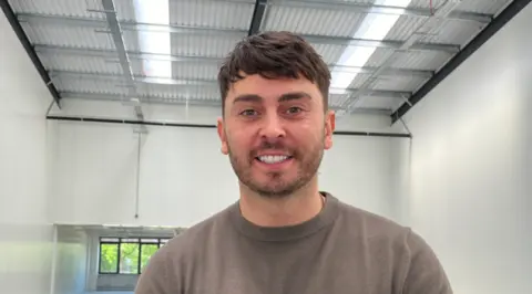 A man in a brown T-shirt with dark hair is smiling at the camera. He is standing inside in a big empty space with white walls and windows in the background beneath a large wall.