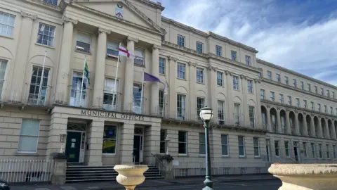 The front of Cheltenham Borough Council's civic offices. The picture shows a four-storey regency building with a sign saying "Municipal Offices". The image has been taking on a dry, cloudy day.
