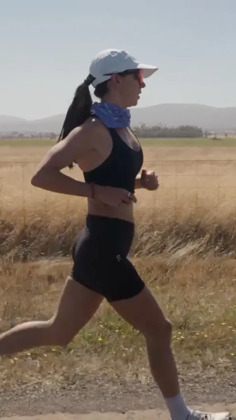 Woman wearing a white hat, neckscarf, sunglasses and a black running top and shorts running past a field