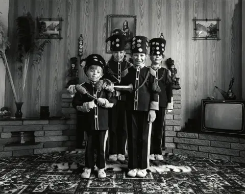 Izabela Jedrzejczyk A black and white photograph of four children standing in a living room dressed in black jazz band uniforms with long, column hats. The uniforms are military-style and the child front-right is holding a small, brass instrument. They are all smiling into the camera and they are standing on a heavily patterned carpet in front of a brick fireplace. The wall above the fireplace has wood panels and a few photographs. There is a television on the right.