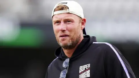 A man with short light brown hair and a beard is wearing a white baseball cap backwards and a dark blue jacket with 'Northern Super Chargers' written on it.