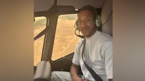 A man in a white shirt sits in a helicopter with a headset on. He smiles at the camera.
