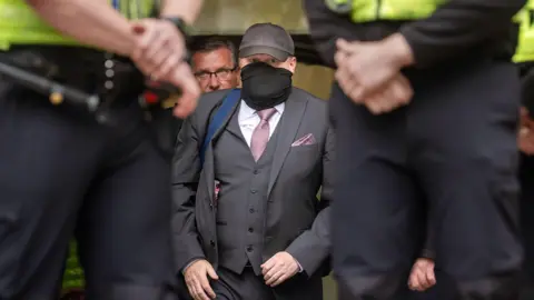 PA Media A man wearing a grey cap and black face covering, grey suit and lilac tie is walking out of Hull Crown Court and is flanked by people, including police officers who are standing in front of him.