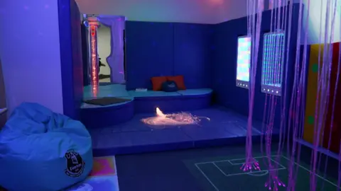 A bean bag and comfortable seating is next to cables with pink lights trailing to the floor. An electronic virtual football game is on the floor. There is a tube filled with pink liquid and bubbles. There is calming lighting.