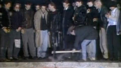 Crowds gather on top of the Berlin Wall as a man uses a pickaxe.